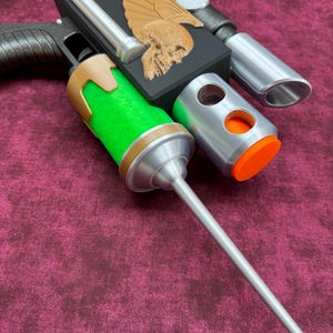May include: A sci-fi prop pistol featuring a black rectangular body with a gold-colored winged skull emblem. It includes a bright green cylindrical section, a long silver needle-like barrel, a silver scope, and an orange circular end cap.