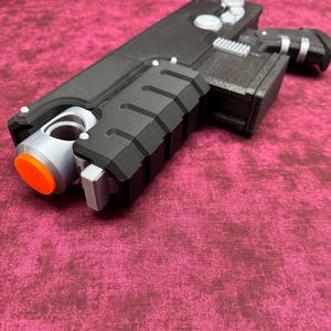 May include: A black and gray toy gun with an orange tip. The toy gun has a rectangular body with a textured grip and several silver-colored buttons and accents. The gun is resting on a maroon surface.