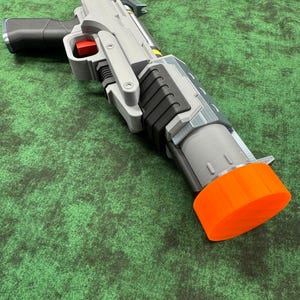 May include: A futuristic toy blaster featuring a grey and black body with a bright orange muzzle cap. The device includes a black pistol grip, a red trigger, and a cylindrical barrel assembly with visible mechanical detailing and layered construction.