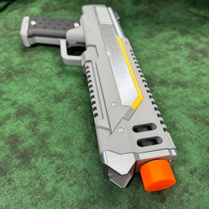 May include: A gray toy pistol featuring a black textured grip, a silver side panel with a yellow accent stripe, and a bright orange muzzle cap. The toy gun has a rectangular barrel design with visible screws and a rail system along the top edge.