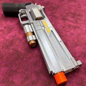 May include: A futuristic silver prop revolver featuring a black textured grip, a cylindrical chamber with visible copper-colored rounds, a mounted brown cylindrical accessory, yellow rectangular accents on the barrel, and a bright orange muzzle tip.