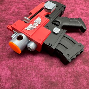 以下が含まれることがあります： 赤と黒のおもちゃの銃で、頭蓋骨と翼のエンブレムが付いています。おもちゃの銃は、黒いハンドルとマガジン、シルバーとオレンジのアクセントが付いています。オブジェクトはマルーンの表面にあります。