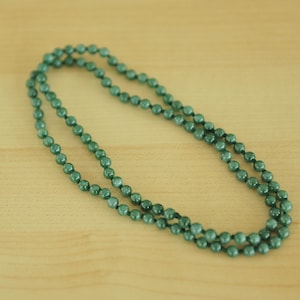 May include: A long necklace composed of small, round, dark green beads. The beads are strung together, forming a double strand that lies on a light brown wooden surface. The necklace is a simple, elegant accessory.