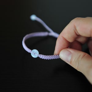 Pode incluir: Uma pulseira de cordão lavanda com uma pequena conta de pedra circular verde-pálida no centro. A pulseira é construída com um padrão de nós trançados e inclui extremidades de cordão ajustáveis com uma pequena conta correspondente na ponta.