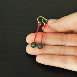 May include: A pair of dangle earrings featuring dark green beads suspended from red cords. The earrings have gold-colored ear wires. The beads have a glossy finish. The earrings are held in a hand against a dark background.