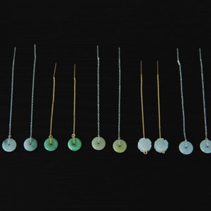 May include: A collection of ten threader earrings arranged in a row. The earrings feature thin silver or gold chains with circular, light green jade-like stone pendants. Some pendants are smooth discs, while others have a carved floral edge detail.