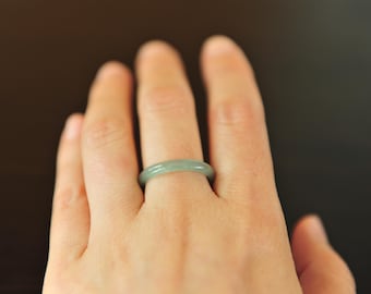 May include: A smooth, solid band ring crafted from pale green jadeite. The ring features a simple, rounded profile with a polished finish, showcasing the natural translucent quality and subtle color variations of the stone material.