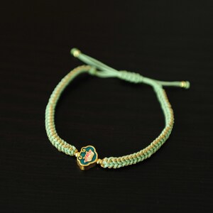 May include: A mint green braided bracelet with gold accents and a gold charm. The charm features a teal background with a pink and gold floral design. The bracelet is adjustable with a sliding knot closure.