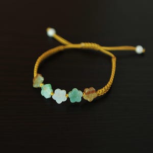 May include: A mustard yellow cord bracelet featuring five small, carved stone flower beads in shades of white, light green, and brown. The bracelet has an adjustable sliding knot closure with two small white beads at the ends of the cord strings.