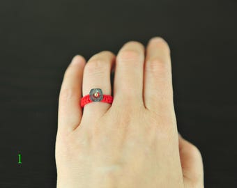 May include: A red macrame ring with a square, translucent, gray-green bead and a small gold-colored bead in the center. The ring is on a hand against a dark background. The ring is a unique piece of jewelry.