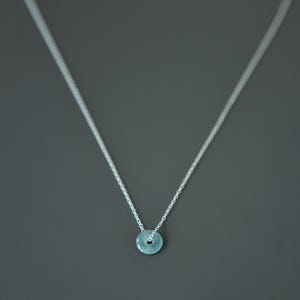 May include: A delicate silver chain necklace featuring a small, circular, pale blue stone pendant with a center hole. The thin metal chain connects to the top of the round, smooth, light blue gemstone, creating a simple and clean jewelry piece.