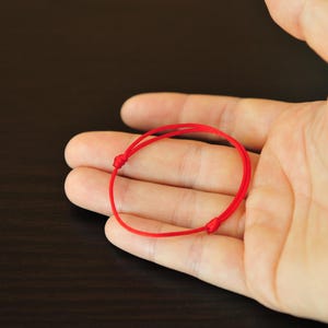 May include: A thin, vibrant red cord bracelet featuring a simple circular design. The bracelet is constructed from a smooth, flexible string material and includes two small, adjustable sliding knots that allow for changes in the circumference.