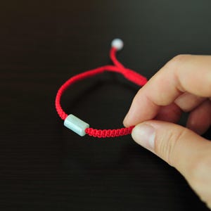 May include: A red braided cord bracelet featuring a single cylindrical pale green stone bead. The bracelet has an adjustable closure mechanism with a small white bead at the end of the cord, designed for a comfortable fit on the wrist.