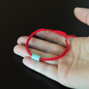 May include: A red cord bracelet featuring a single cylindrical light green stone bead. The bracelet is constructed with a braided cord design and includes an adjustable sliding knot mechanism to secure the fit around the wrist.
