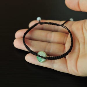 May include: A bracelet featuring a dark brown braided cord with an adjustable sliding knot closure. A small, circular, light green jade-like stone bead with a central hole is centered on the cord, accented by two tiny white beads at the ends.