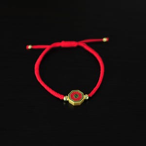 May include: Red string bracelet with a gold-colored, octagonal charm. The charm has a red and green design. The bracelet is tied with a sliding knot for adjustability. The background is black.