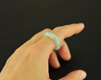 May include: A light green jade ring is worn on a finger. The ring has a smooth, polished surface and a rounded shape. The jade has a translucent quality, with subtle variations in color.