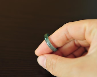 May include: A smooth, solid band ring crafted from translucent dark green jade stone. The ring features a polished, rounded surface with subtle variations in color intensity, showcasing the natural depth and texture of the polished mineral material.
