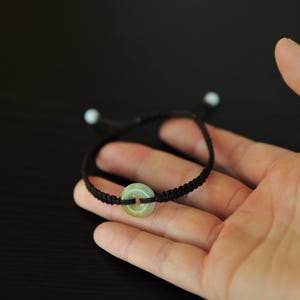 May include: A bracelet featuring a dark brown woven cord and a central light green circular stone bead with a hole in the middle. The cord has two smaller light green beads at the ends of the adjustable pull strings, creating a simple design.