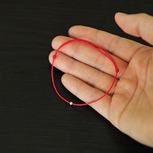 May include: A simple red string bracelet with a small, silver-colored bead. The bracelet is knotted and rests on a person's open palm against a dark background. The bracelet is a minimalist design.