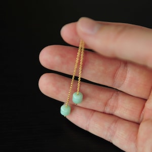 Op de afbeelding: Een paar delicate gouden kettingoorbellen met lichtgroene jade kralen aan de uiteinden.