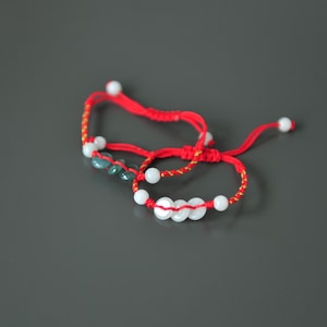 May include: Two red string bracelets with white and green beads. One bracelet has white beads and the other has green beads.