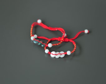 May include: Two red string bracelets with white and green beads. One bracelet has white beads and the other has green beads.