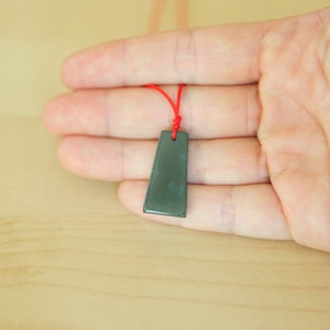 May include: A dark gray pendant with a rectangular shape hangs from a red string. The pendant is smooth and has a slight sheen.