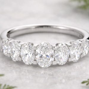 May include: A silver band ring featuring seven oval-cut diamonds. The diamonds are set closely together, creating a continuous sparkle. The ring is displayed on a light-colored surface, with greenery in the background.