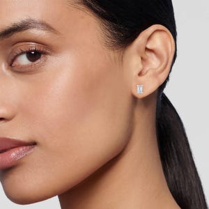May include: Close-up of a person's ear with a small, rectangular-cut diamond stud earring. The earring is set in a silver or white gold setting. The skin tone is light, and the background is a neutral gray.