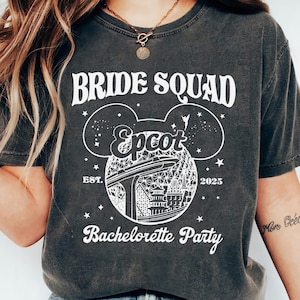 Op de afbeelding: Donkergrijs t-shirt met witte tekst en afbeeldingen. Het shirt toont "BRIDE SQUAD" boven een Mickey Mouse-hoofdontwerp met "Epcot" en "Bachelorette Party" eronder. Het jaar "2025" staat ook op het shirt.