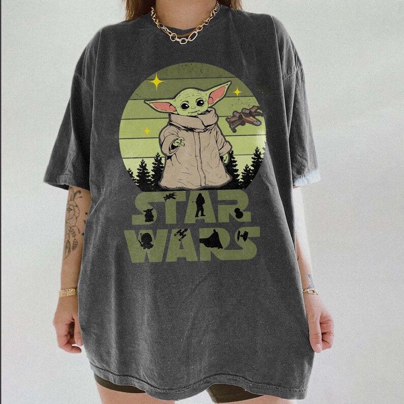 Pode incluir: Camiseta cinza escuro com um gr&aacute;fico de Grogu de Star Wars. O design inclui Grogu em um casaco bege, uma nave espacial e as palavras "STAR WARS" em verde. A camiseta tem mangas curtas e um corte folgado.