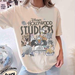 May include: Ivory-colored t-shirt featuring a graphic of Disney's Hollywood Studios characters, including Mickey Mouse, Minnie Mouse, Donald Duck, and Goofy. The design incorporates the park's logo and various icons. The shirt also has the text "Comfort Colors" and "Ivory".