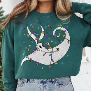 May include: A teal green sweatshirt featuring a white cartoon ghost-like character wrapped in colorful Christmas lights. The character holds a candy cane and a small pumpkin. Gold stars and snowflakes are scattered around the design.