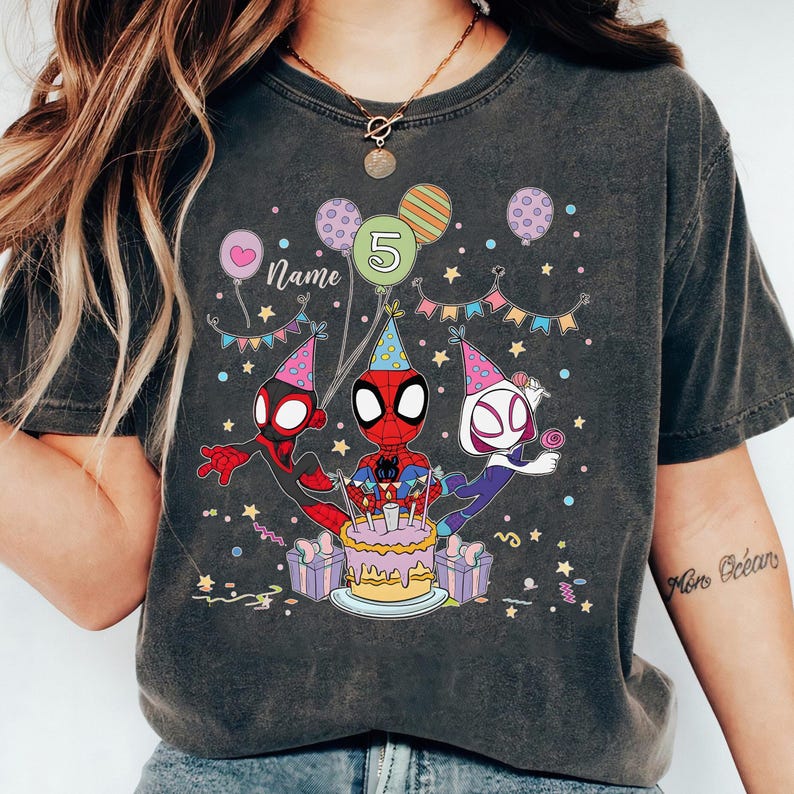 May include: Dark gray t-shirt featuring a colorful graphic of Spider-Man and friends celebrating a birthday. The design includes a cake, balloons, and the number 5. The word "Name" is also visible.