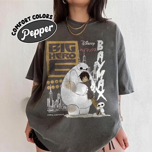 May include: Dark grey t-shirt featuring a graphic from the Disney movie "Big Hero 6". The design includes Baymax hugging Hiro Hamada, with the words "Big Hero 6" and "Baymax" in white and gold. The shirt also has the text "Comfort Colors Pepper".