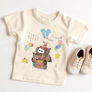 May include: Cream-colored toddler t-shirt featuring a cartoon image of Tow Mater from the movie Cars, wearing a party hat and surrounded by balloons and a birthday cake. A pair of beige canvas sneakers are next to the shirt.
