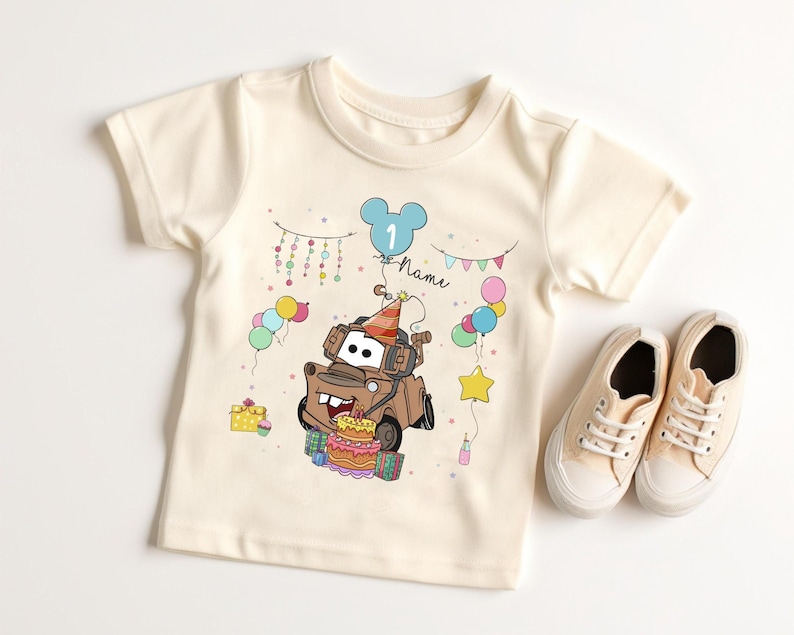 May include: Cream-colored toddler t-shirt featuring a cartoon image of Tow Mater from the movie Cars, wearing a party hat and surrounded by balloons and a birthday cake. A pair of beige canvas sneakers are next to the shirt.