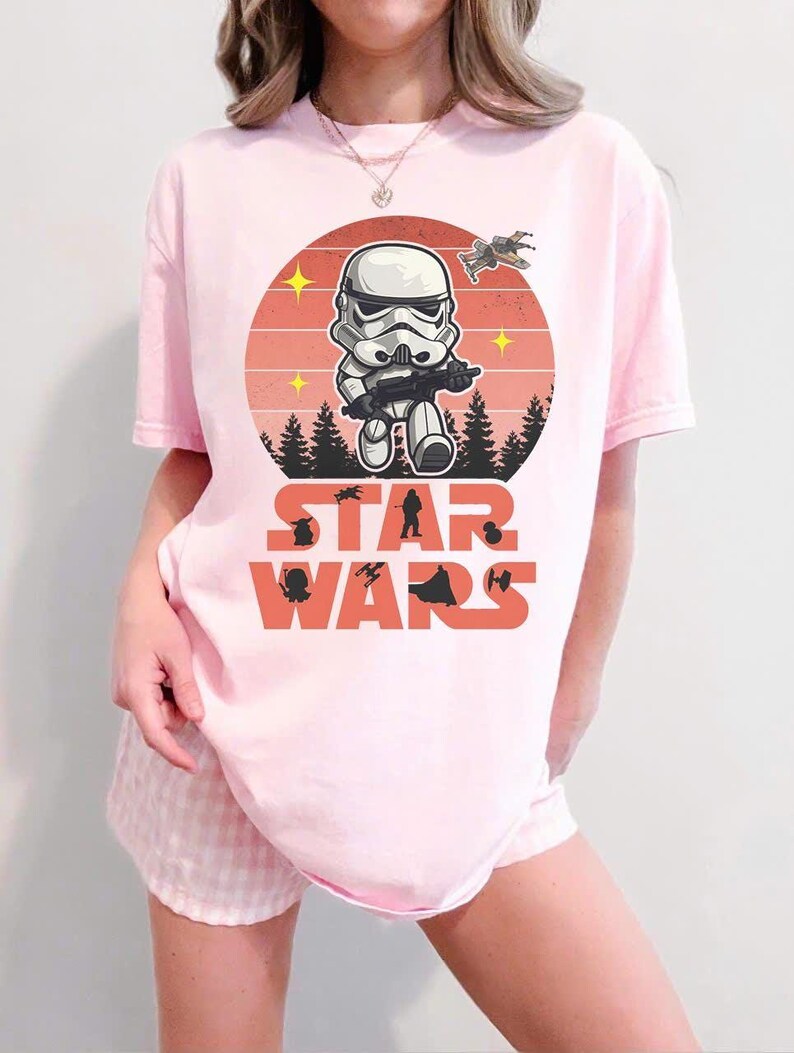 Pode incluir: Camiseta rosa claro com um design de Stormtrooper de desenho animado segurando um blaster, em um c&iacute;rculo com cores de p&ocirc;r do sol com o texto "STAR WARS" abaixo. Inclui silhuetas de &aacute;rvores e naves espaciais.
