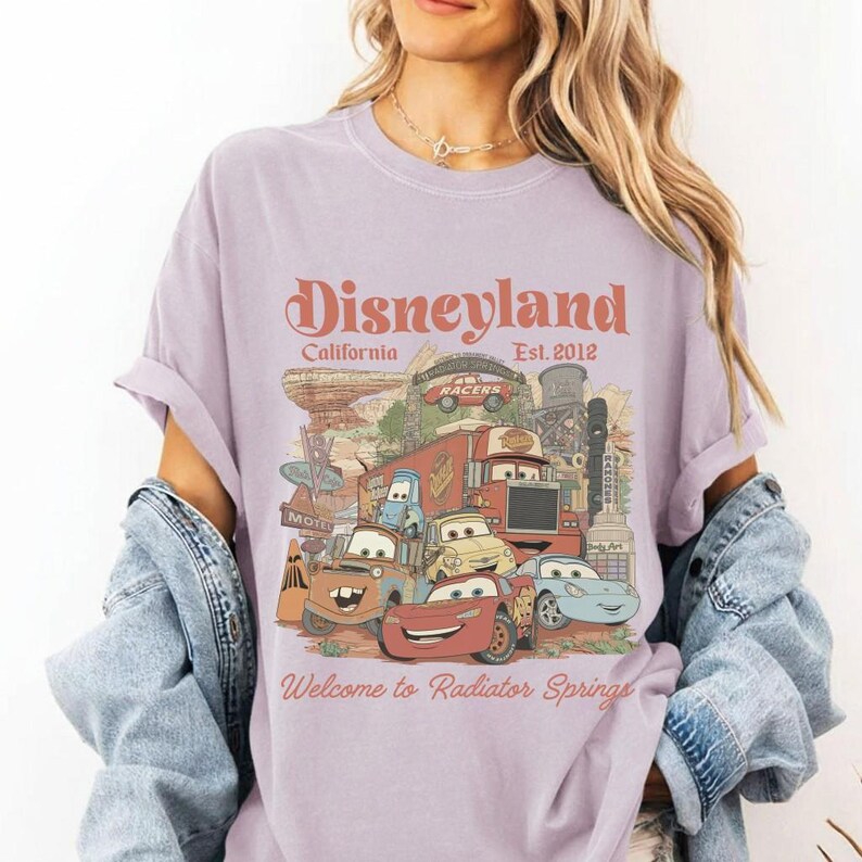 May include: A lilac-colored t-shirt featuring a graphic of Disneyland and the text "Welcome to Radiator Springs." The design includes illustrations of animated cars and architectural elements. The text "Disneyland California Est. 2012" is also visible.
