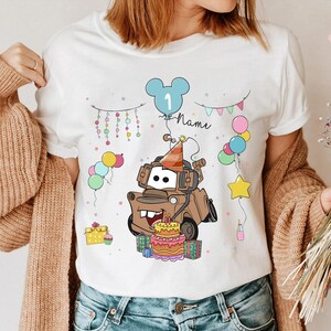 May include: White t-shirt featuring a cartoon image of Tow Mater from the movie Cars, wearing a party hat and surrounded by balloons and a birthday cake. The number 1 and the word "Name" are also included in the design.
