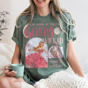 May include: Green t-shirt with a graphic featuring the words "Glinda Wicked" in pink, along with illustrations of a princess and a crown. The shirt also has the text "You're Gonna Be Popular" and "Toss Toss".