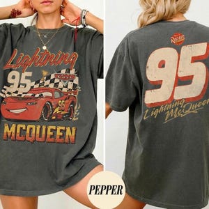 May include: Dark gray t-shirt featuring a graphic of Lightning McQueen from the Cars movie. The front displays the number 95, the character's name, and a checkered flag. The back of the shirt also features the number 95 and the Lightning McQueen name.