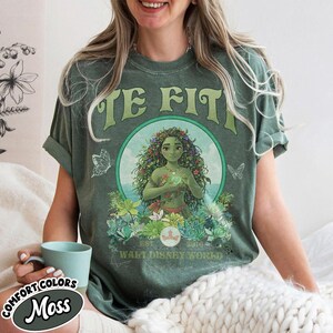 May include: A moss green t-shirt with a graphic of a woman surrounded by flowers and the text "TE FIT". The shirt also features the text "WALT DISNEY WORLD" and "COMFORT COLORS".