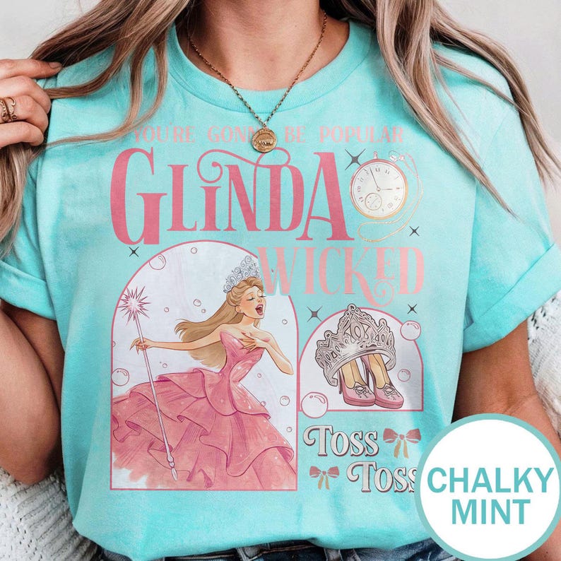 May include: A light blue t-shirt with a graphic of Glinda from Wicked, featuring the text "You're Gonna Be Popular" and "Glinda Wicked Toss Toss". The design includes a pink dress, crown, and clock.