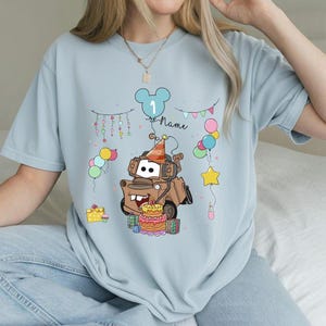 May include: Light blue t-shirt featuring a cartoon illustration of a brown truck wearing a party hat, with a birthday cake and balloons. The design includes the number "1" and the word "Name". Colorful bunting and gift boxes complete the festive design.
