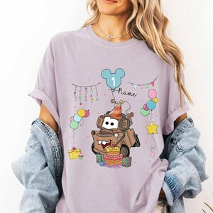 May include: A light purple t-shirt featuring a cartoon illustration of a brown tow truck with a birthday cake, balloons, and the number 1. The design includes the word "Name" and festive decorations, perfect for a first birthday celebration.