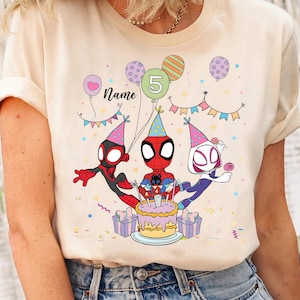 May include: A cream-colored t-shirt featuring a colorful birthday design with Spider-Man characters. The graphic includes a birthday cake, balloons, and the number 5. The word "Name" is also visible.