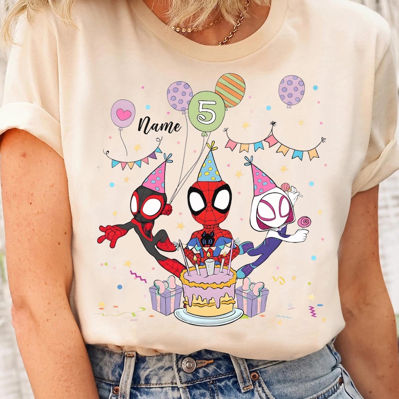 May include: A cream-colored t-shirt featuring a colorful birthday design with Spider-Man characters. The graphic includes a birthday cake, balloons, and the number 5. The word "Name" is also visible.