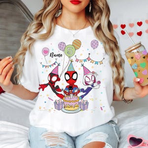 May include: White t-shirt featuring a colorful graphic of Spider-Man characters celebrating a birthday. The design includes balloons, a cake, and the number 5. The word "Name" is also included in the design.